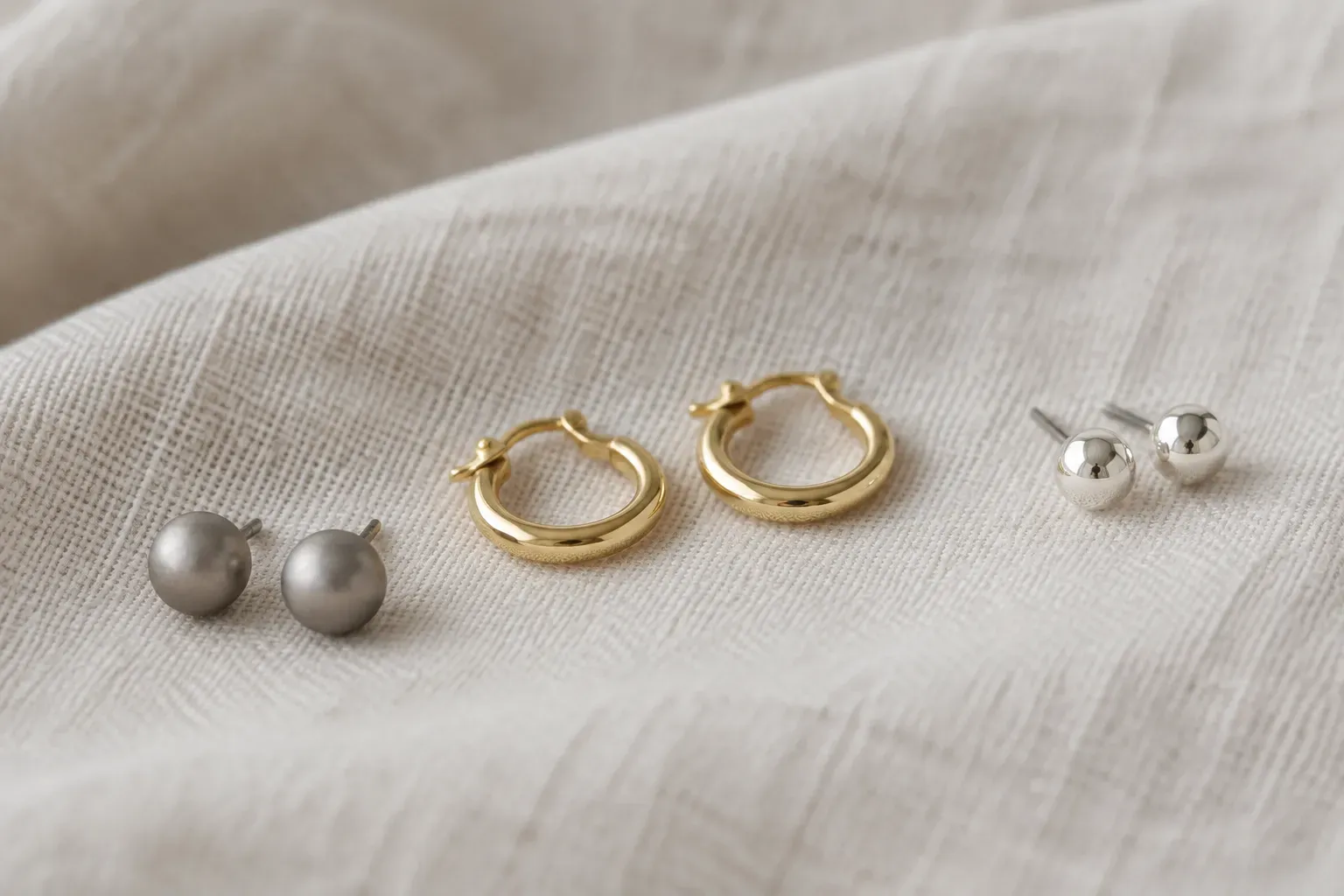 Titanium ball studs, small gold hoops and sterling silver studs resting on white linen