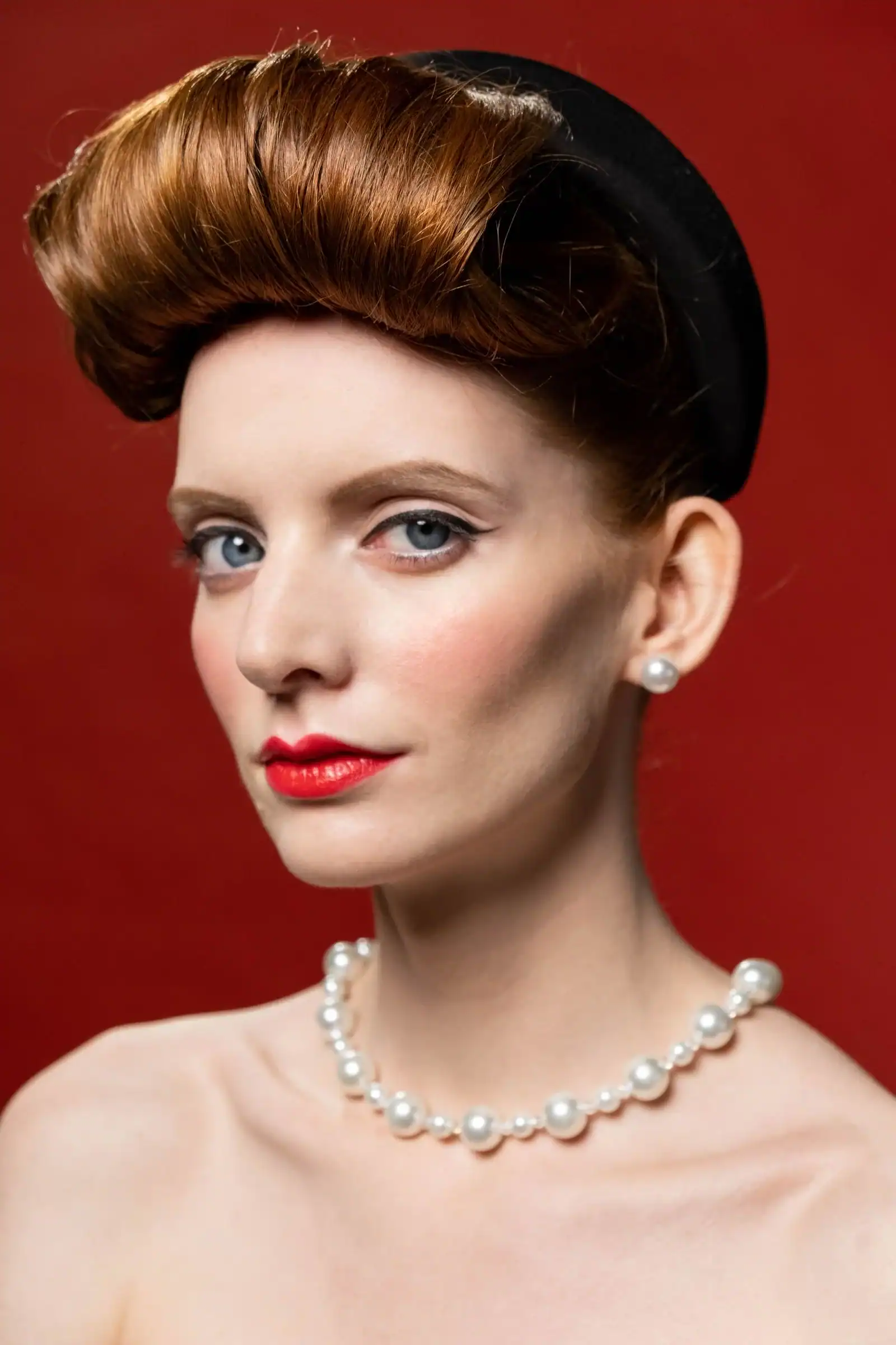 A woman with auburn victory rolls and a pearl necklace against a red wall