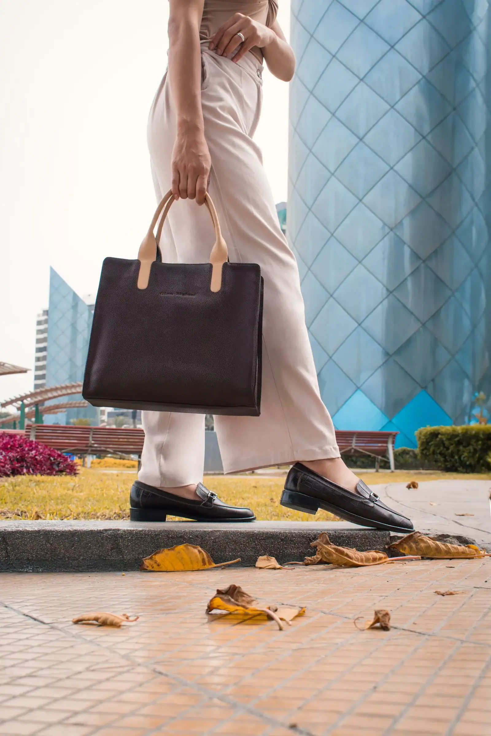 A woman in cream trousers and loafers carrying a dark leather bag